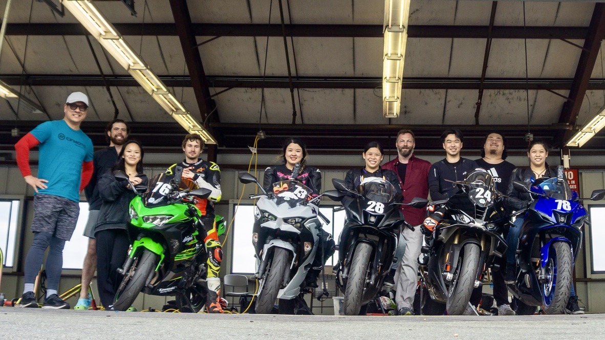 A group of riders standing with their motorcycles in the paddock.